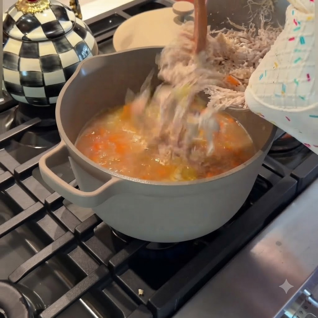 Versamento di pollo finemente sfilacciato da una ciotola di metallo in una pentola grigia di zuppa di pollo e verdure in ebollizione.