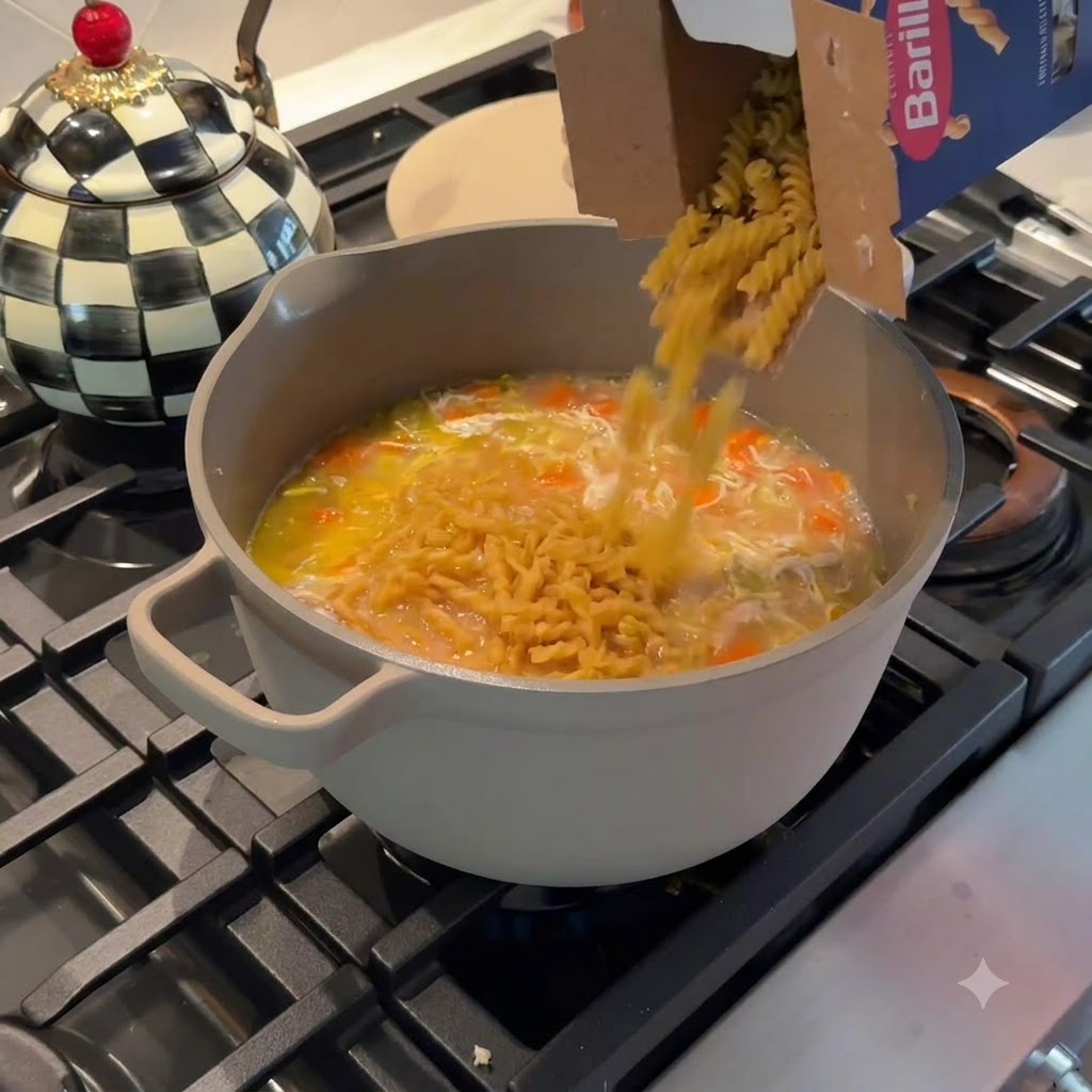 Pasta a spirale secca versata direttamente da una scatola Barilla in una pentola di zuppa di pollo e noodle in ebollizione.