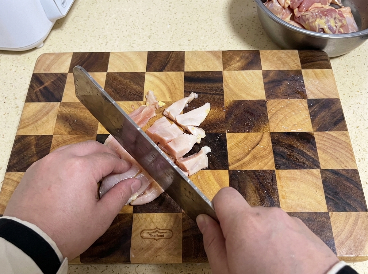 Una mannaia che affetta il petto di pollo crudo in piccoli pezzi su un tagliere di legno a quadri.