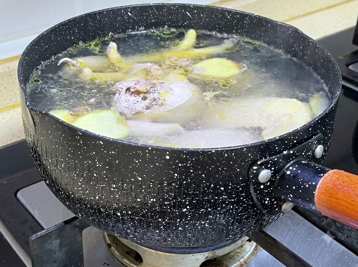 Pezzi di pollo crudo e fette di zenzero che sobbollono in una pentola nera marezzata piena di acqua bollente.