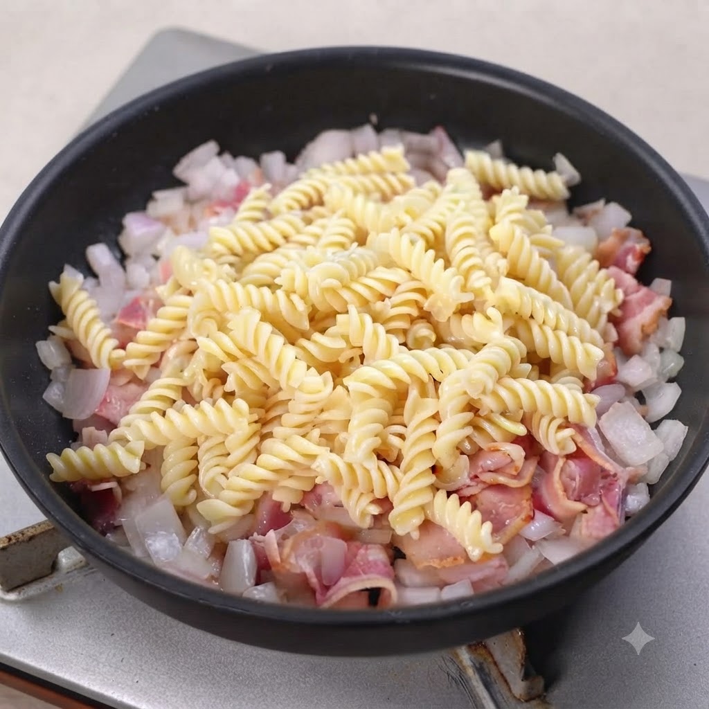 Fusilli cotti adagiati sopra pancetta e cipolle in una padella nera.