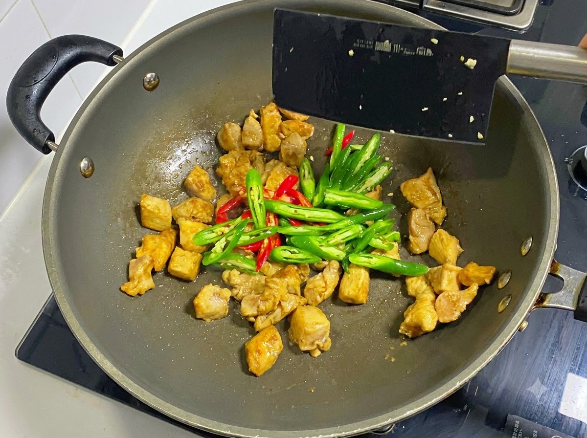 Peperoncini verdi e rossi affettati aggiunti ai pezzi di pollo cotti in un wok e mescolati con una spatola nera.