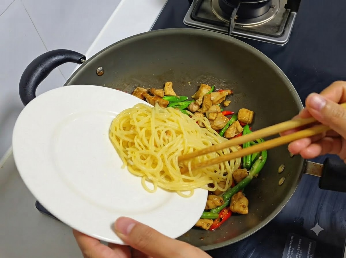 Spaghetti cotti trasferiti da un piatto bianco in un wok contenente pollo e peperoncini cotti.