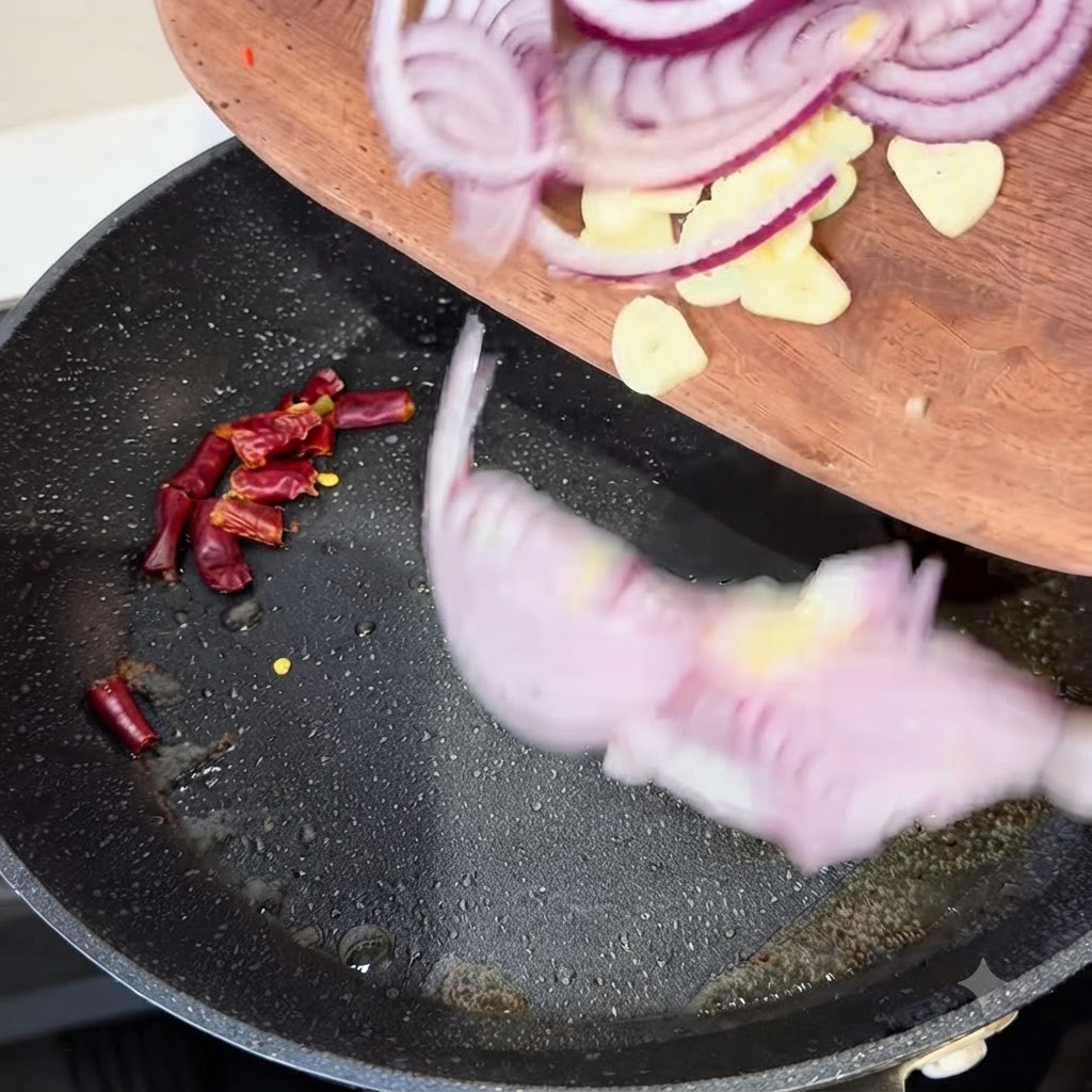 Fette di cipolla rossa, spicchi daglio e peperoncini secchi trasferiti da un tagliere di legno in una padella calda.