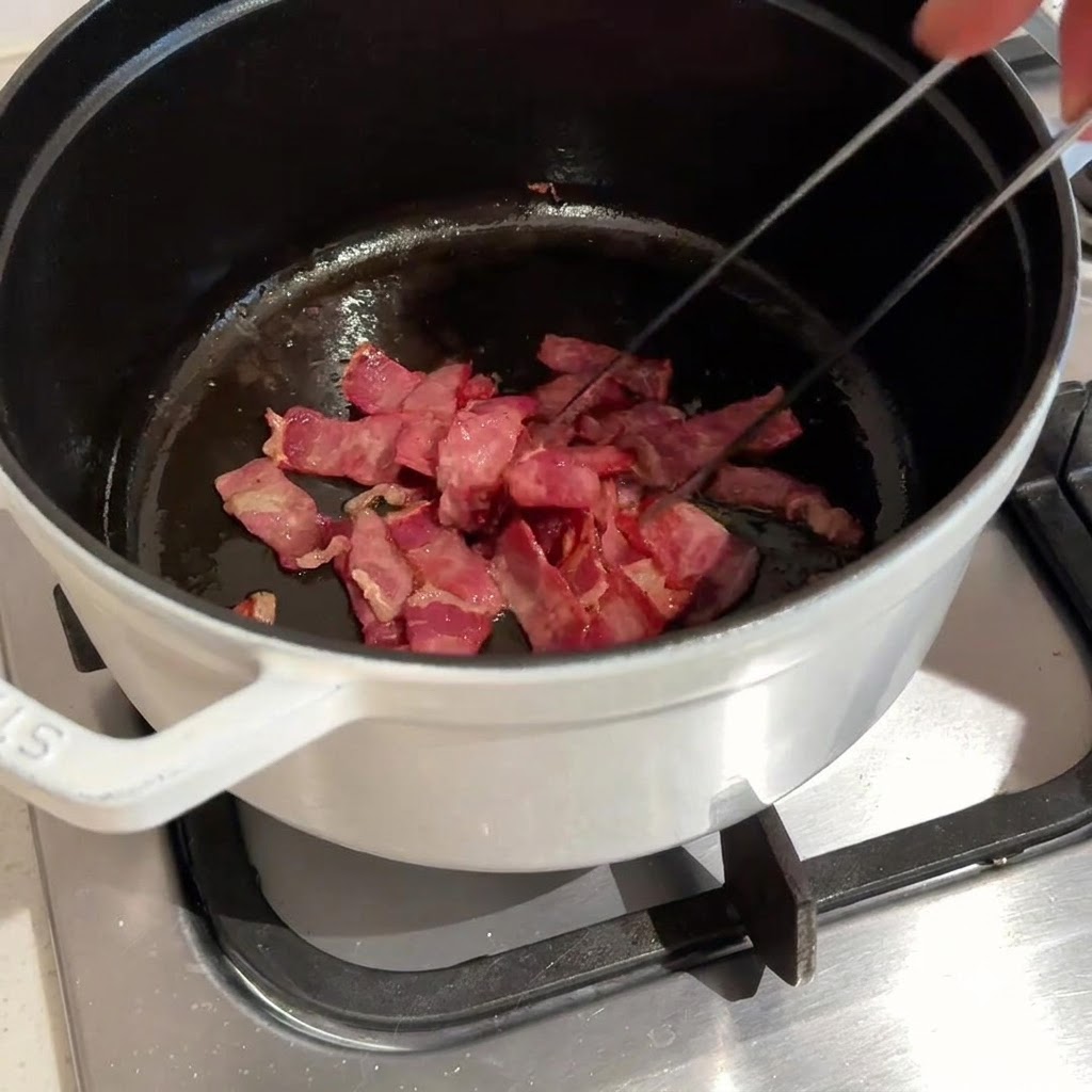 Pancetta tagliata a cubetti saltata e fritta in una pentola scura usando pinze di metallo.