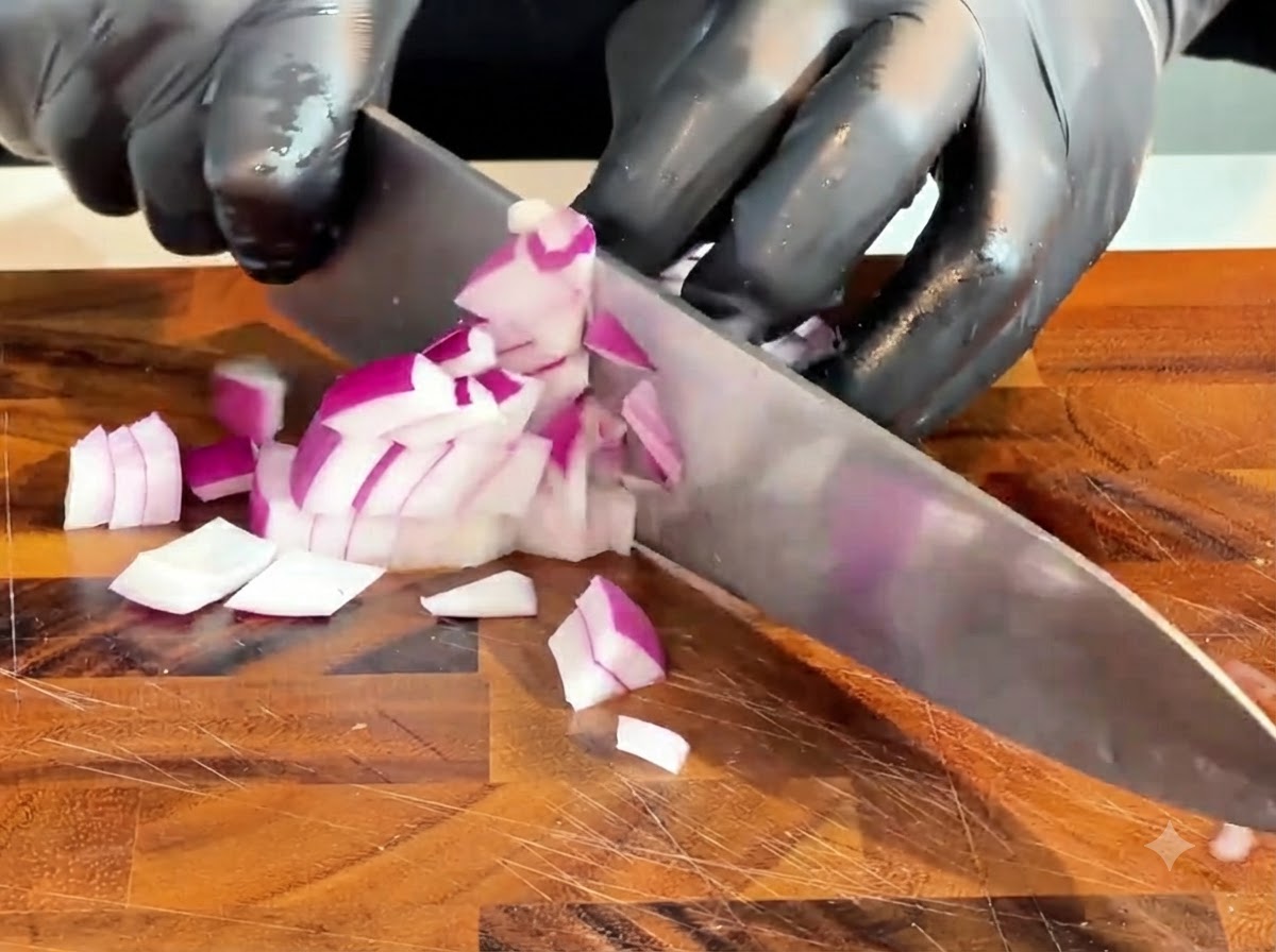 Un coltello che taglia una cipolla rossa cruda a piccoli pezzi su un tagliere di legno.