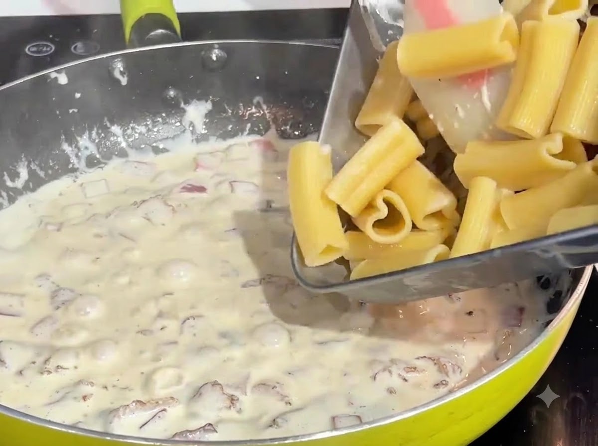 Pasta a forma di cuore precedentemente cotta versata da un vassoio in una salsa di panna e pancetta che bolle in padella.
