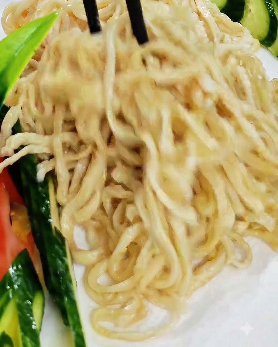 Bacchette nere che sollevano e dispongono i noodles cotti su un piatto bianco guarnito con cetriolo.