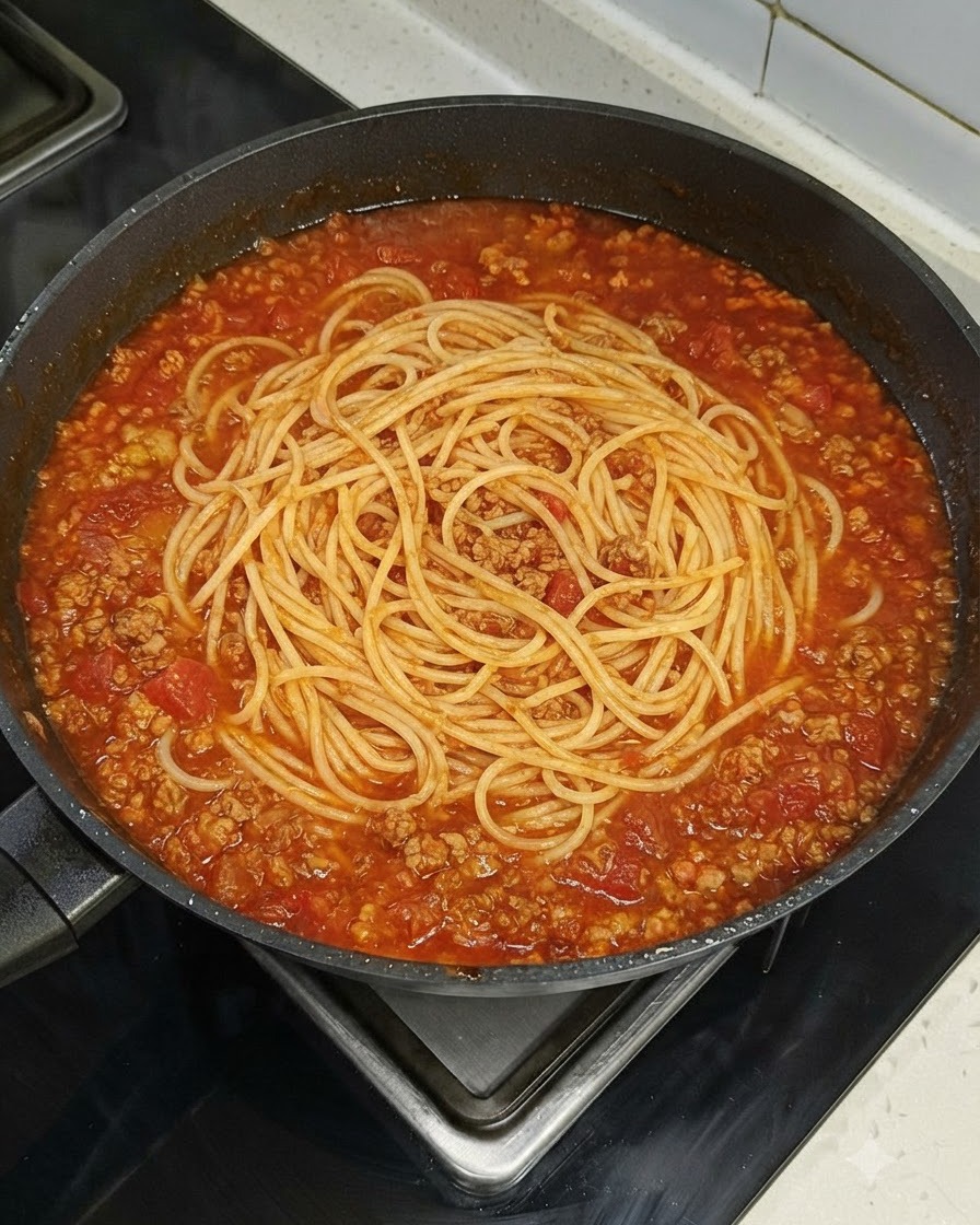 Un nido di spaghetti adagiato uniformemente al centro di una grande padella colma di ragù rosso su un fornello.