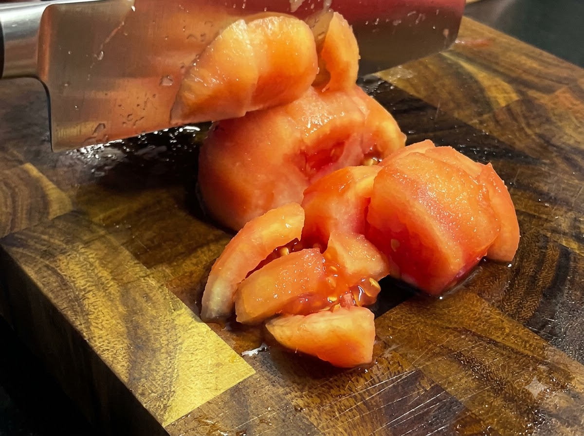 Coltello che taglia i pomodori spellati e cotti a pezzetti su un tagliere di legno.