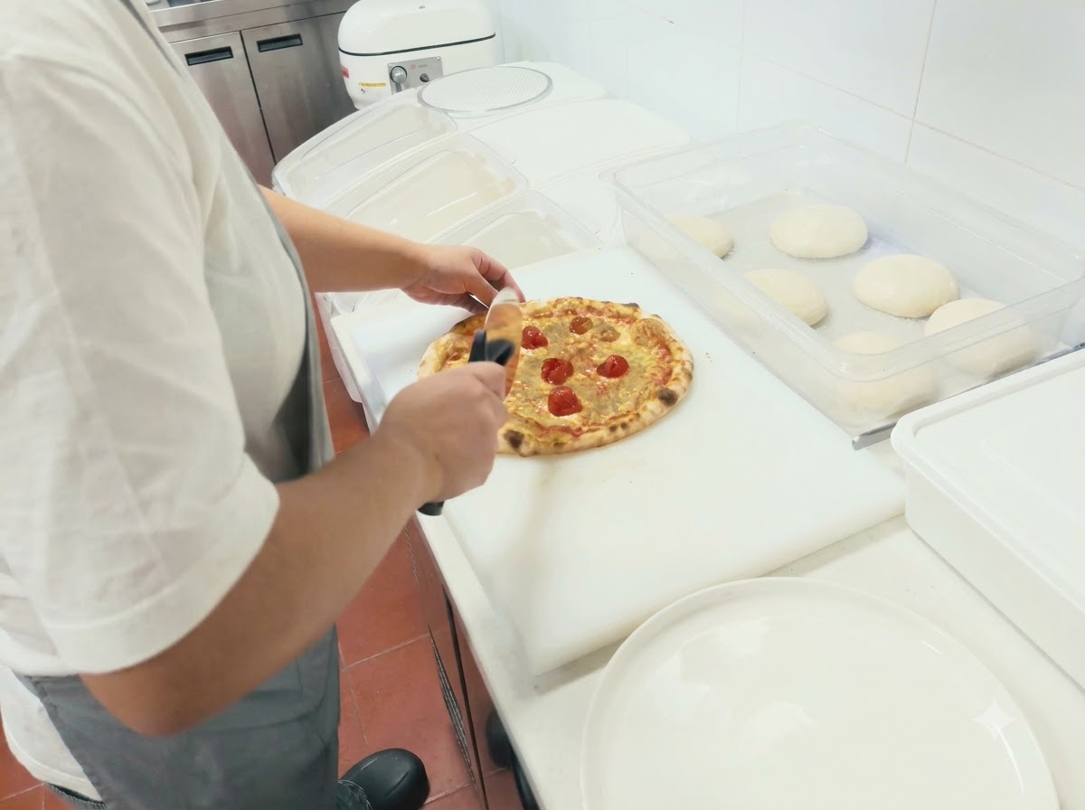 Una persona che usa una rotella tagliapizza per tagliare una pizza appena sfornata su un tagliere bianco.
