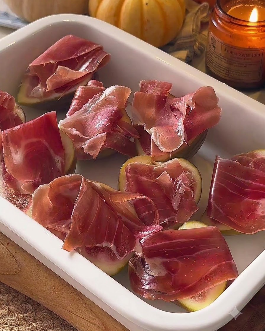Fichi assemblati guarniti con prosciutto disposti in una pirofila bianca.