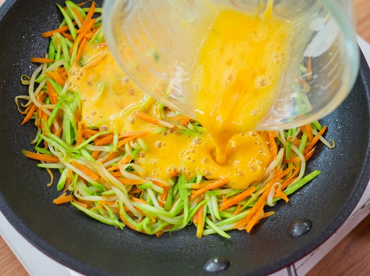 Liquido duovo giallo versato da un misurino sopra lo strato di verdure nella padella.