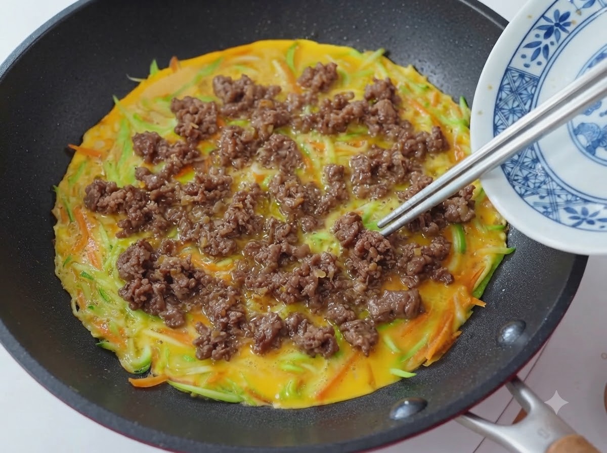 Primo piano di bacchette che posizionano e distribuiscono pezzi di manzo cotto sopra una padella con base di pancake di verdure e uova.