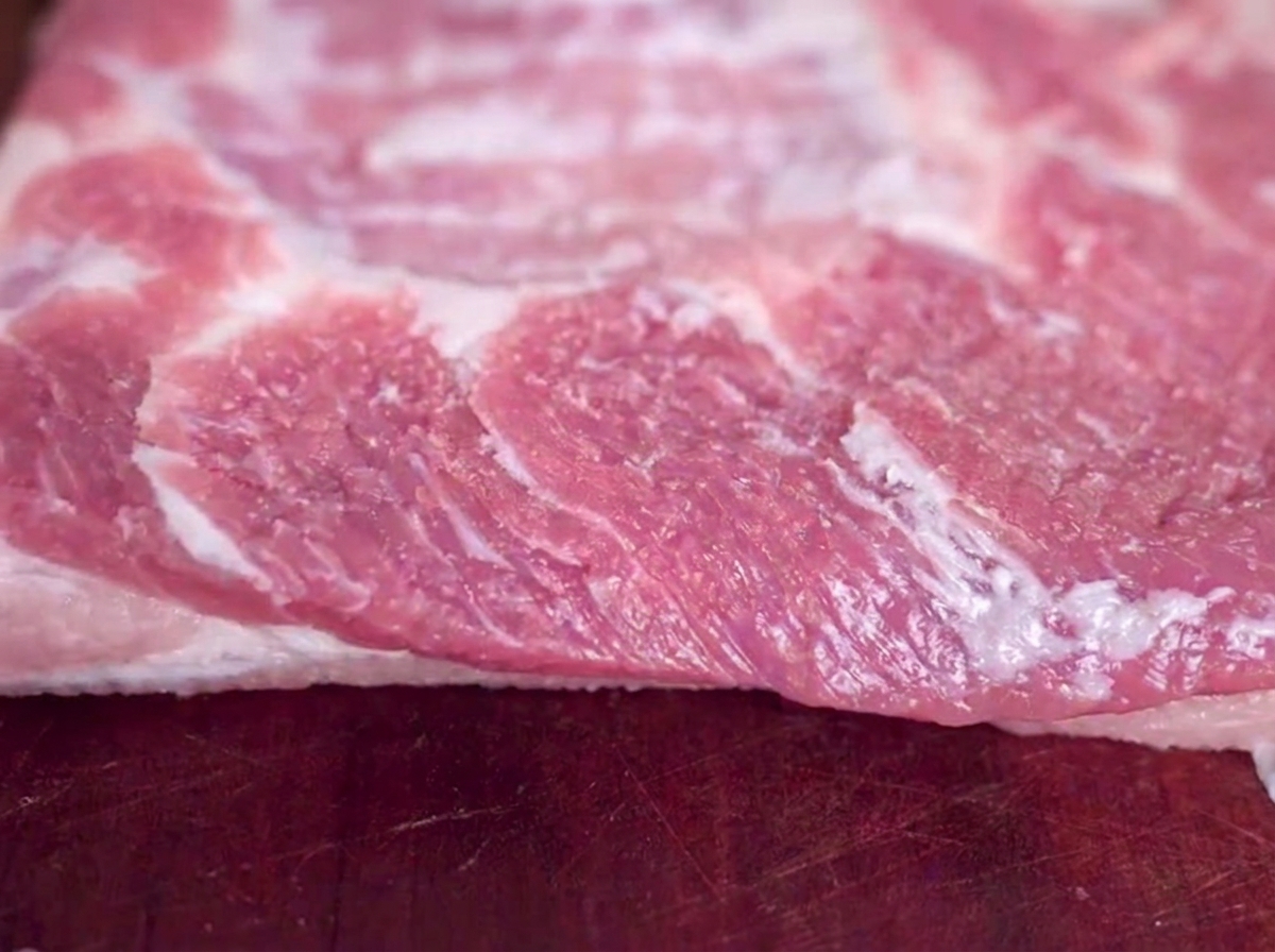 Primo piano di un pezzo crudo di pancetta di maiale su un tagliere di legno con granelli di sale visibili.