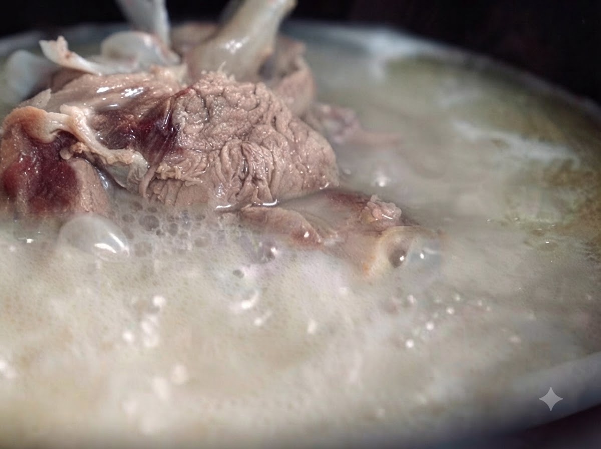 Un brodo di ossa di maiale denso, lattiginoso e opaco che bolle vigorosamente in una pentola, con pezzi di carne e ossa visibili.