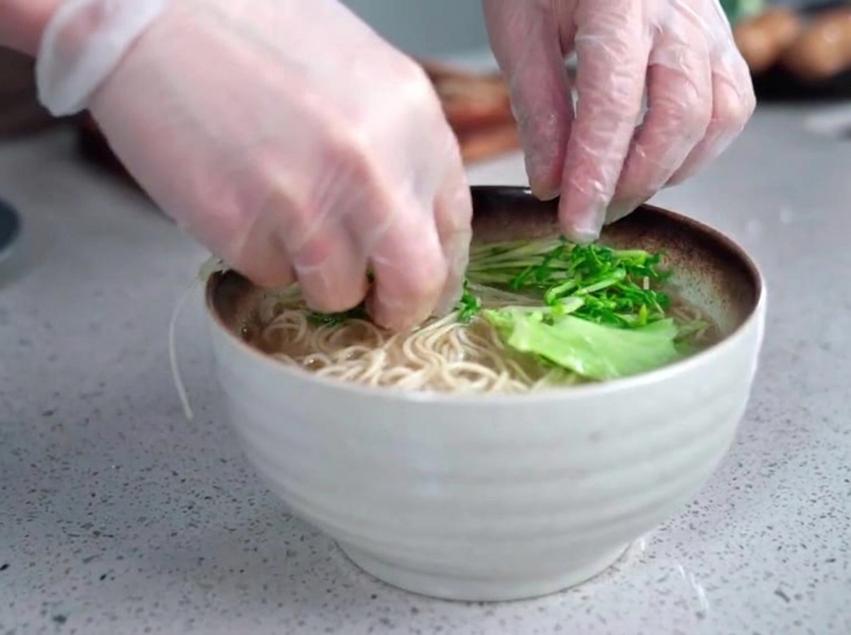 Mani con i guanti che posizionano delicatamente verdure fresche a foglia verde in una ciotola fumante di ramen e brodo opaco.