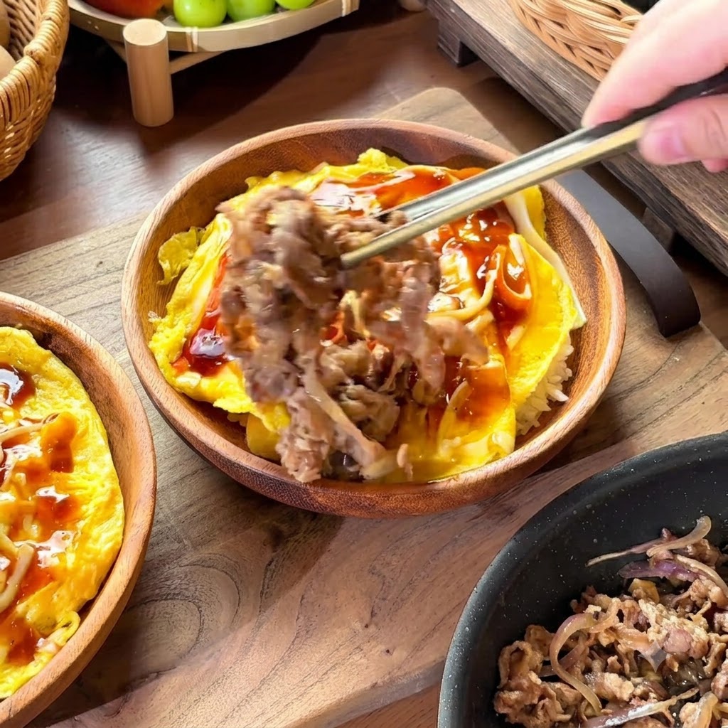 Bacchette che sollevano una porzione di manzo e cipolle cotti per posizionarli su una ciotola di riso e uova coperte di salsa.