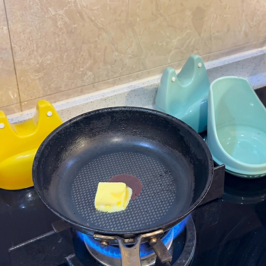 Un cubo di burro giallo che si scioglie al centro di una padella antiaderente nera sopra una fiamma a gas.