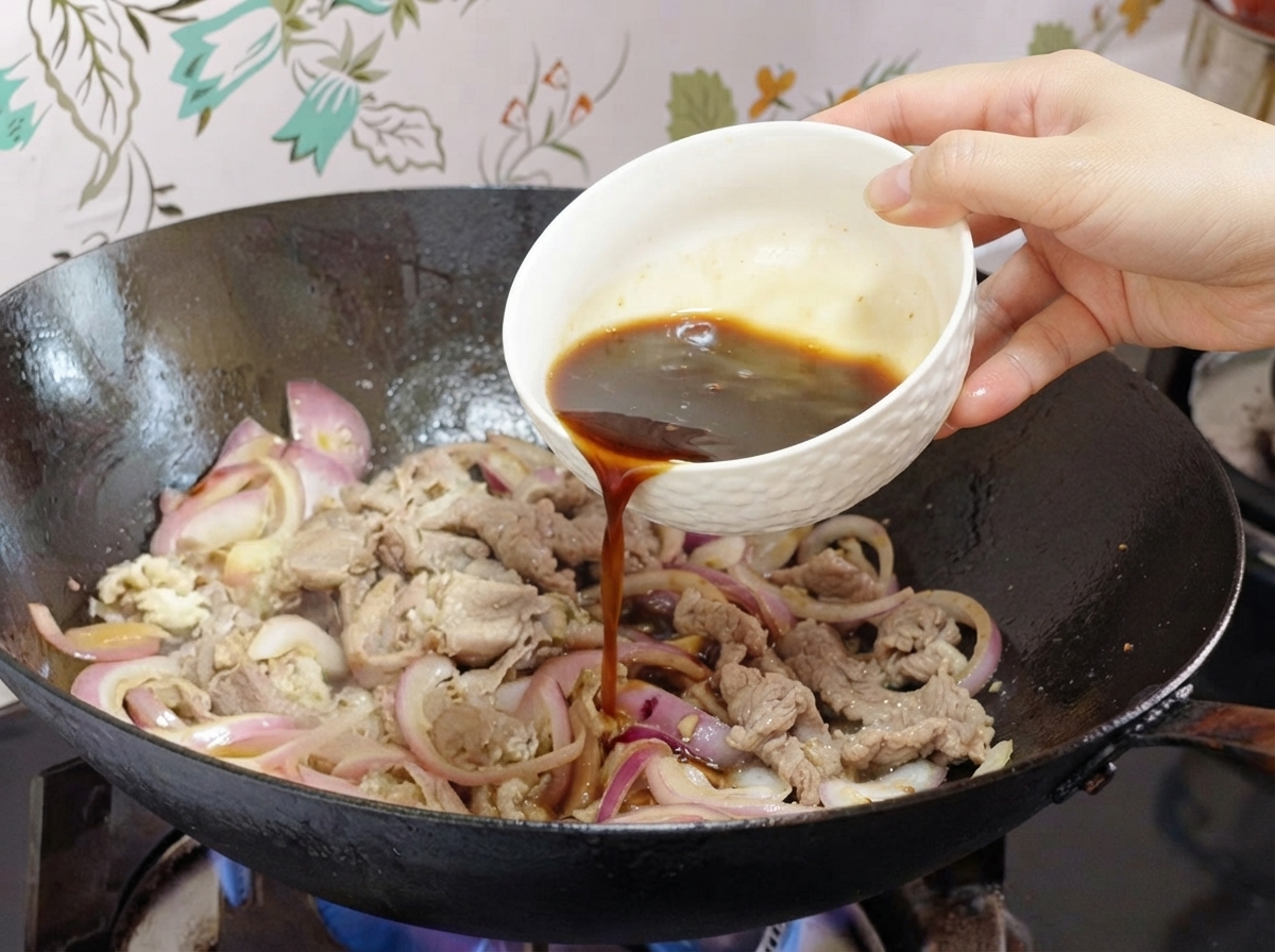 Salsa versata da una piccola ciotola bianca in un wok con cipolle e fette di manzo.