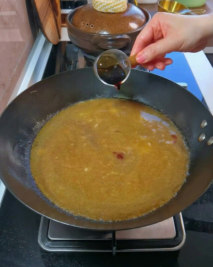 Una mano che versa salsa di soia scura da un piccolo misurino in un wok pieno di brodo.