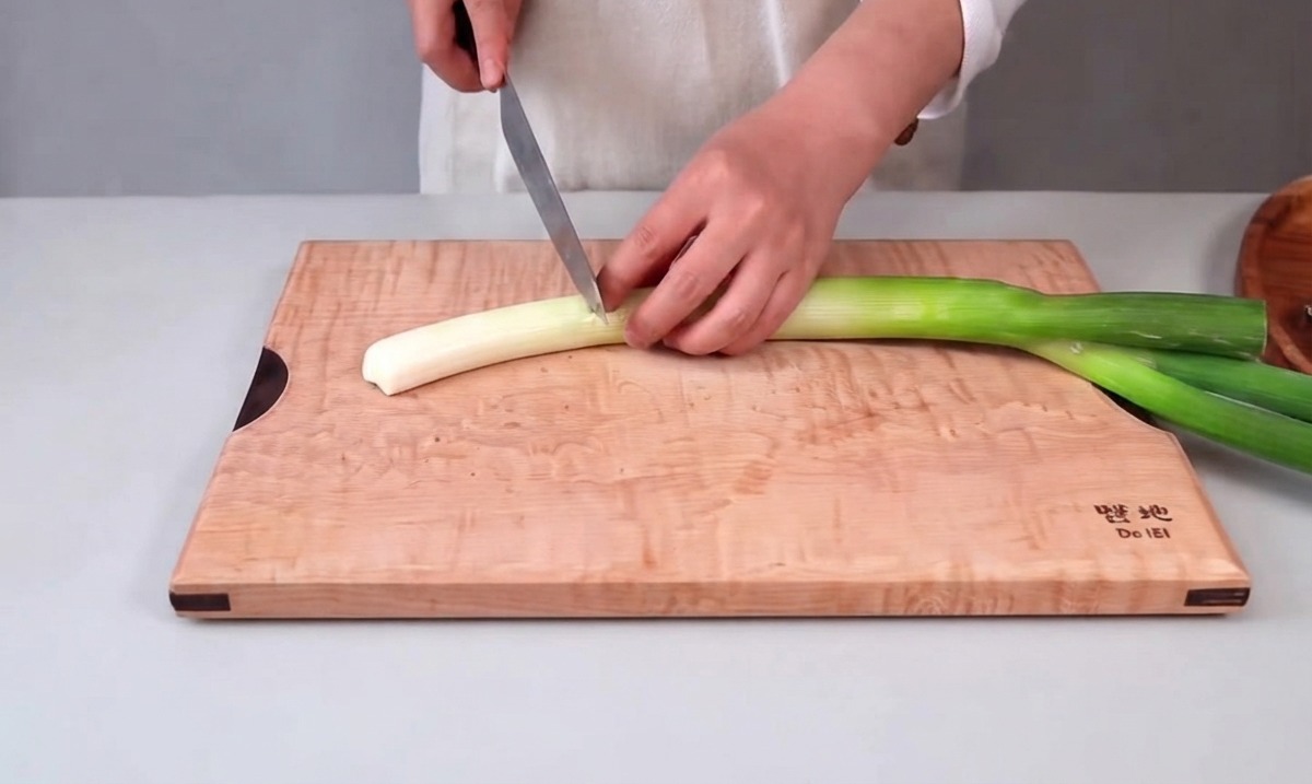 Mani che tengono un coltello da cucina mentre tagliano un grosso cipollotto su un tagliere di legno.