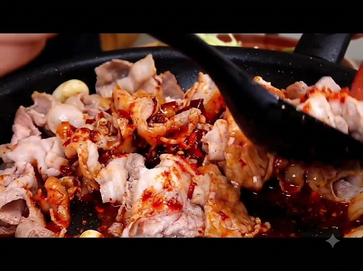 Una spatola nera che mescola e amalgama la salsa rossa piccante con le fette di pancetta di maiale in una padella calda.