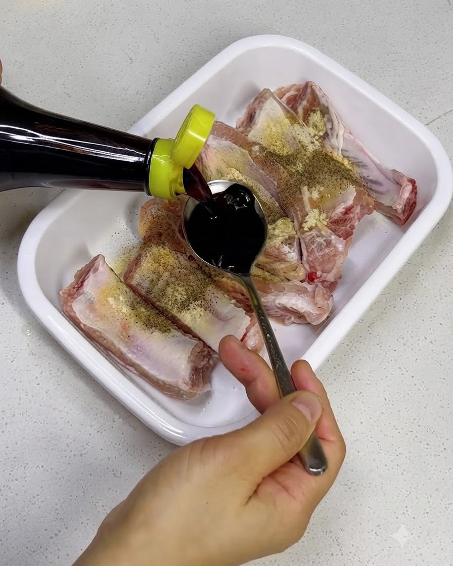Versamento di salsa di soia scura da una bottiglia su costine di maiale crude in un vassoio bianco.