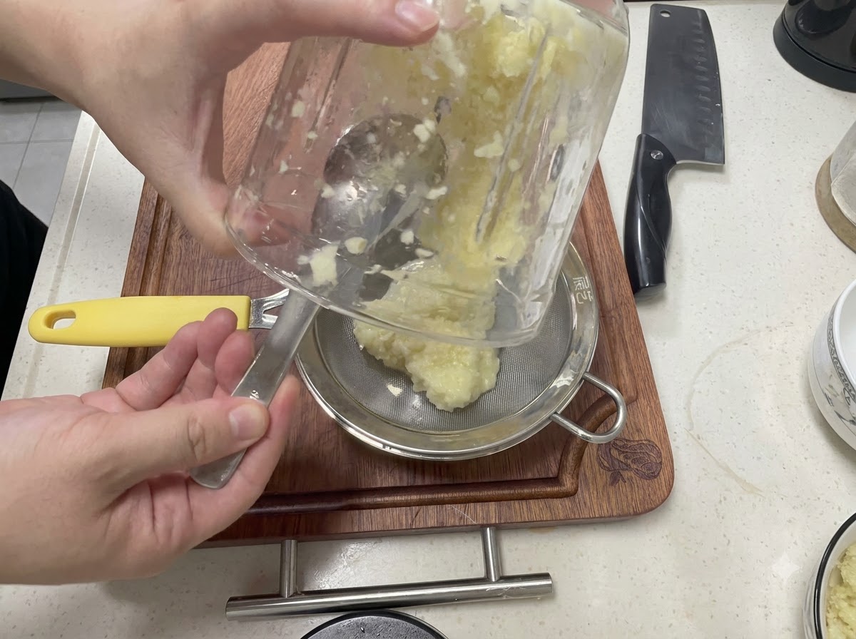 Aglio frullato versato da un contenitore di vetro in un colino a maglia fine sopra una ciotola di metallo.