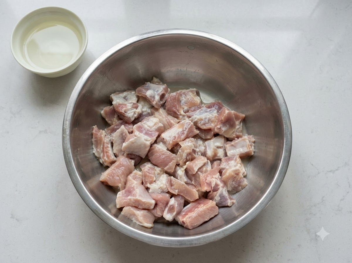 Una ciotola di costine di maiale marinate con una piccola ciotola separata di albume duovo posizionata nelle vicinanze su un piano di lavoro bianco.