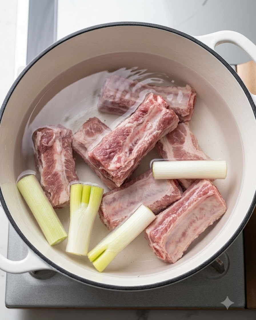 Costine di maiale fresche adagiate in una pentola di acqua fredda con cipollotti e aromi prima dellebollizione.