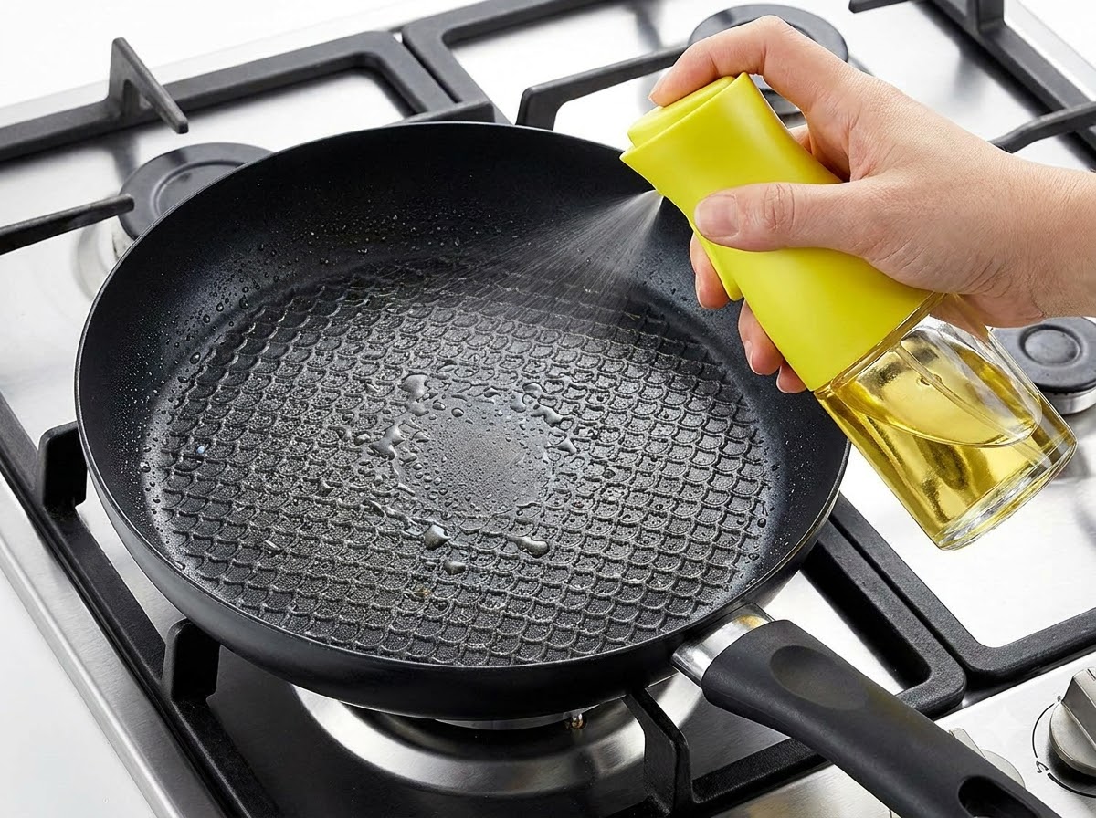 Una mano che spruzza olio da cucina in una padella nera posizionata su un fornello.