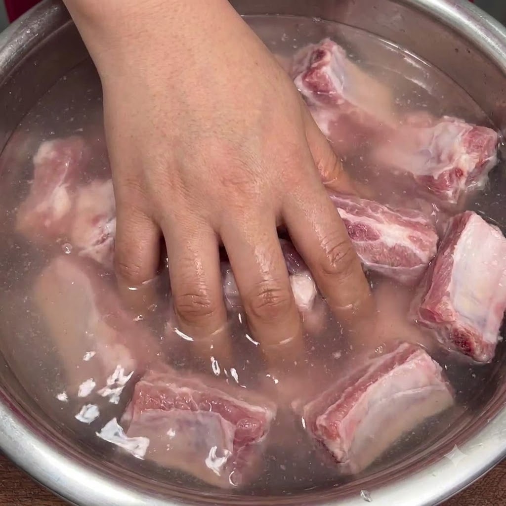 Una mano che mescola energicamente le costine di maiale crude in una ciotola di acqua torbida per distribuire il bicarbonato.