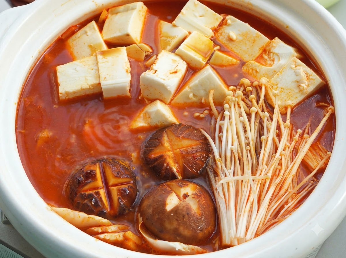 Un hot pot in ceramica bianca con base di zuppa rossa in ebollizione, cubetti di tofu vellutato, funghi shiitake intagliati a croce e funghi enoki sotto un coperchio.