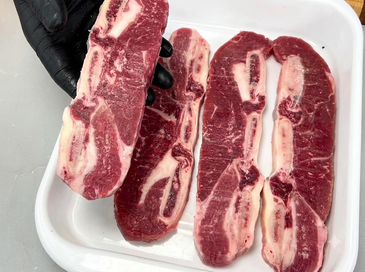 Una mano con i guanti che solleva una costina di manzo LA cruda con osso, mostrando la ricca marmorizzazione e la struttura ossea.