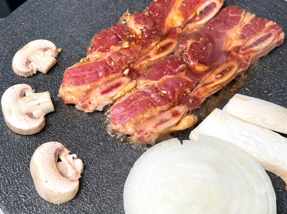 Costine di manzo marinate che sfrigolano su una piastra nera calda accanto a cipolle bianche affettate e funghi.