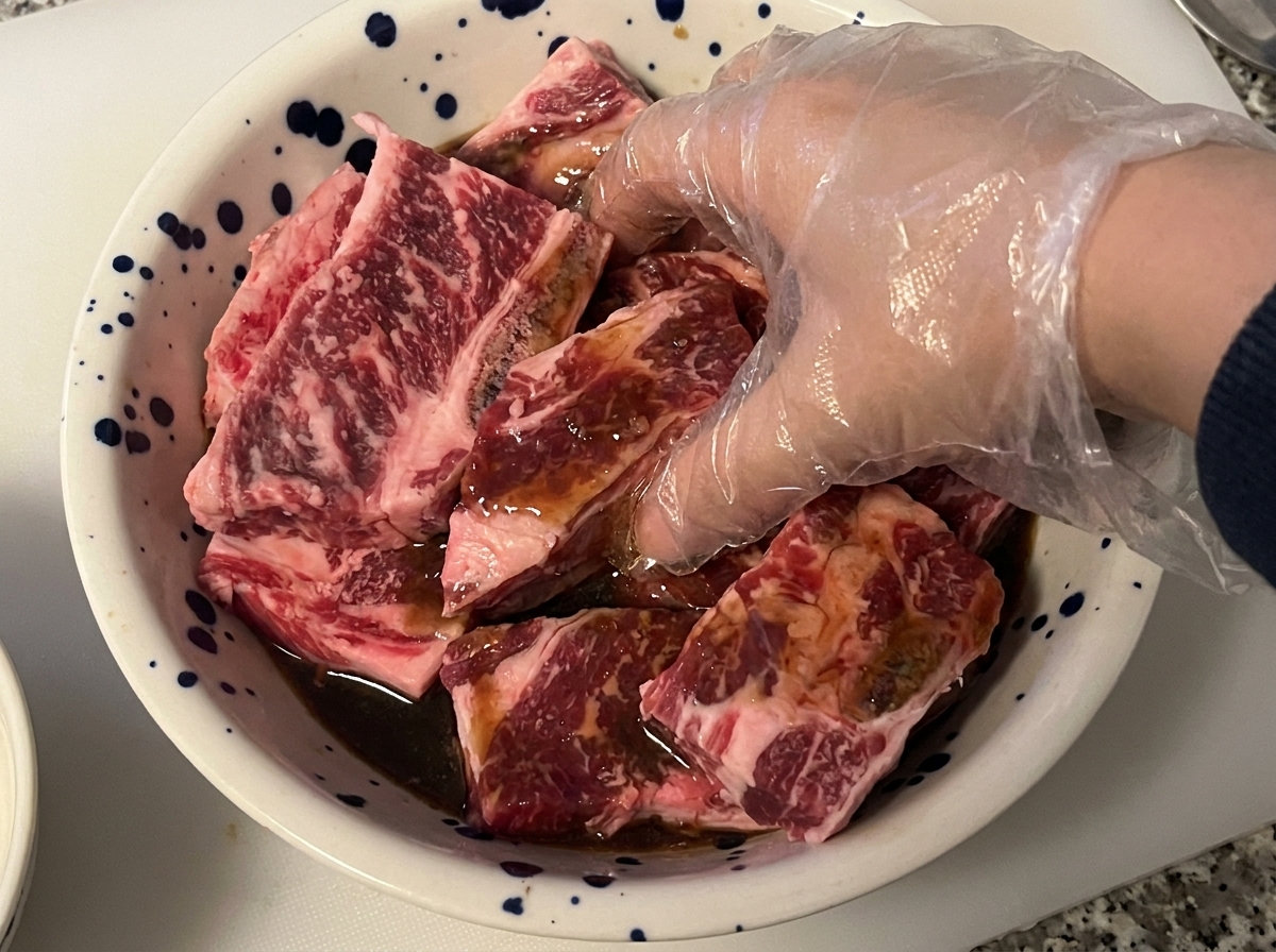 Una mano guantata che mescola e massaggia le costine di manzo crude con una marinata scura in una ciotola.
