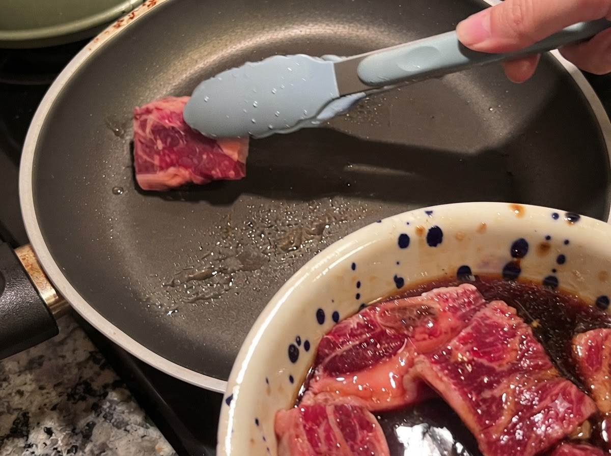 Delle pinze che posizionano un pezzo di costina di manzo marinata in una padella calda.