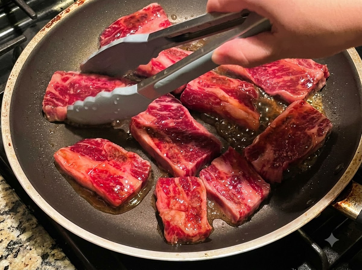 Delle pinze che girano più pezzi di costine di manzo che friggono in una padella.