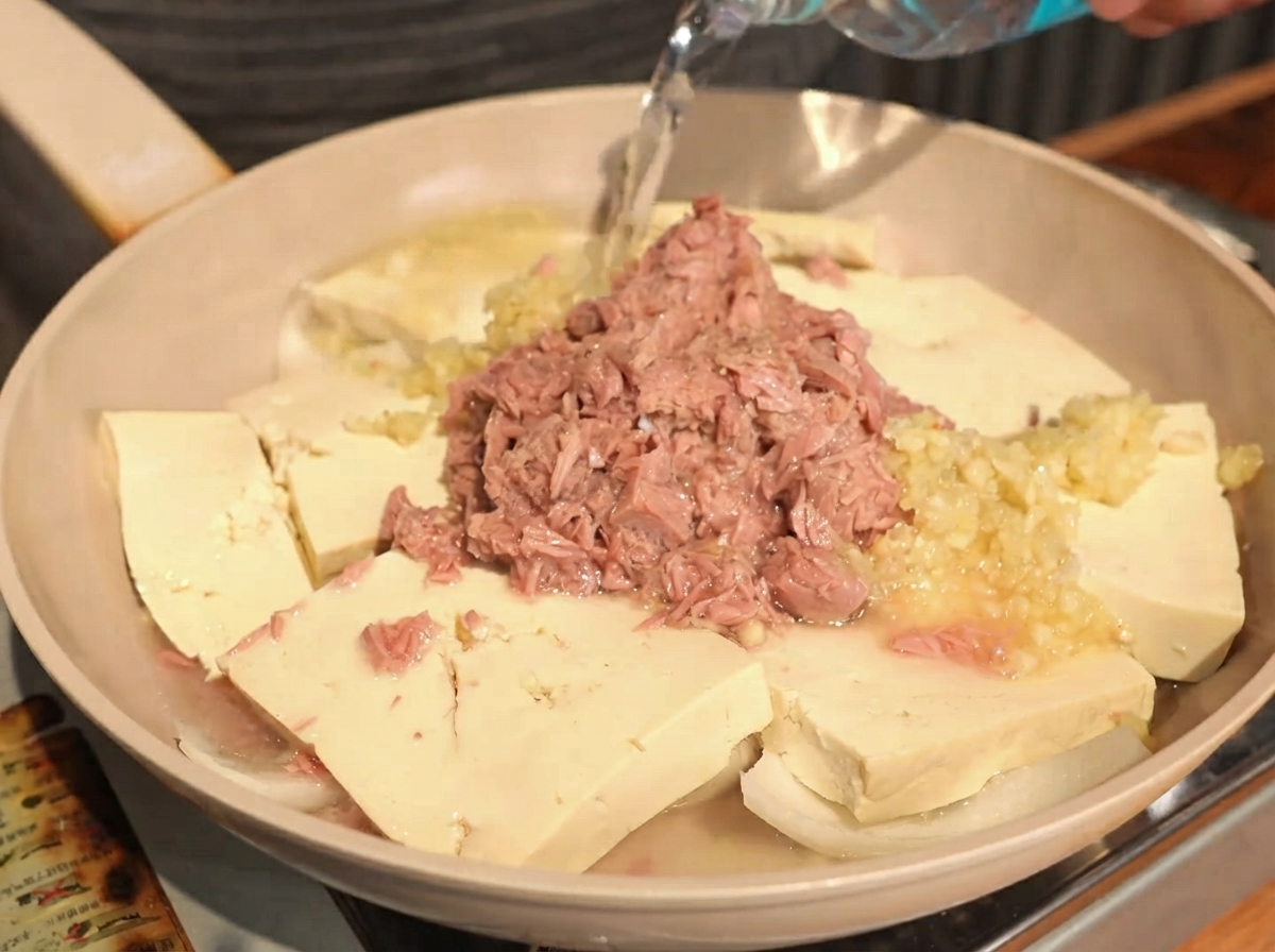 Acqua limpida versata da una bottiglia in una padella contenente tofu, cipolle, aglio e tonno in scatola a strati.