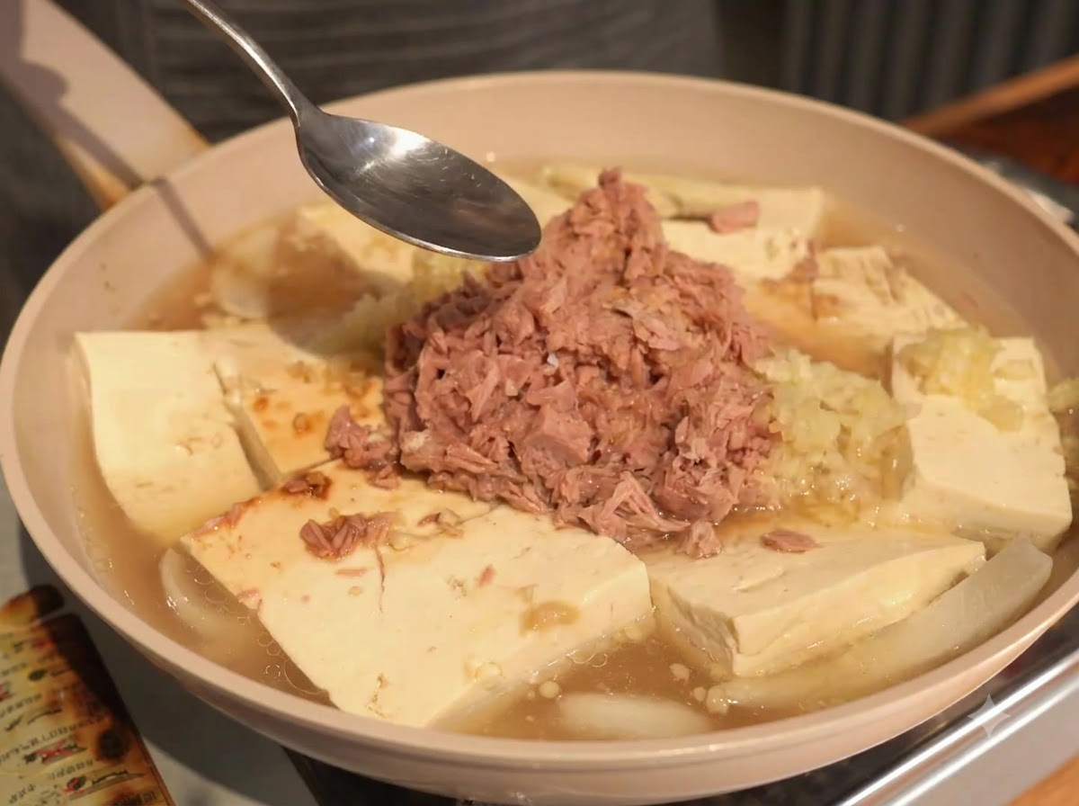 Un cucchiaio tenuto sopra la padella contenente tonno, tofu e brodo liquido durante il processo di condimento.