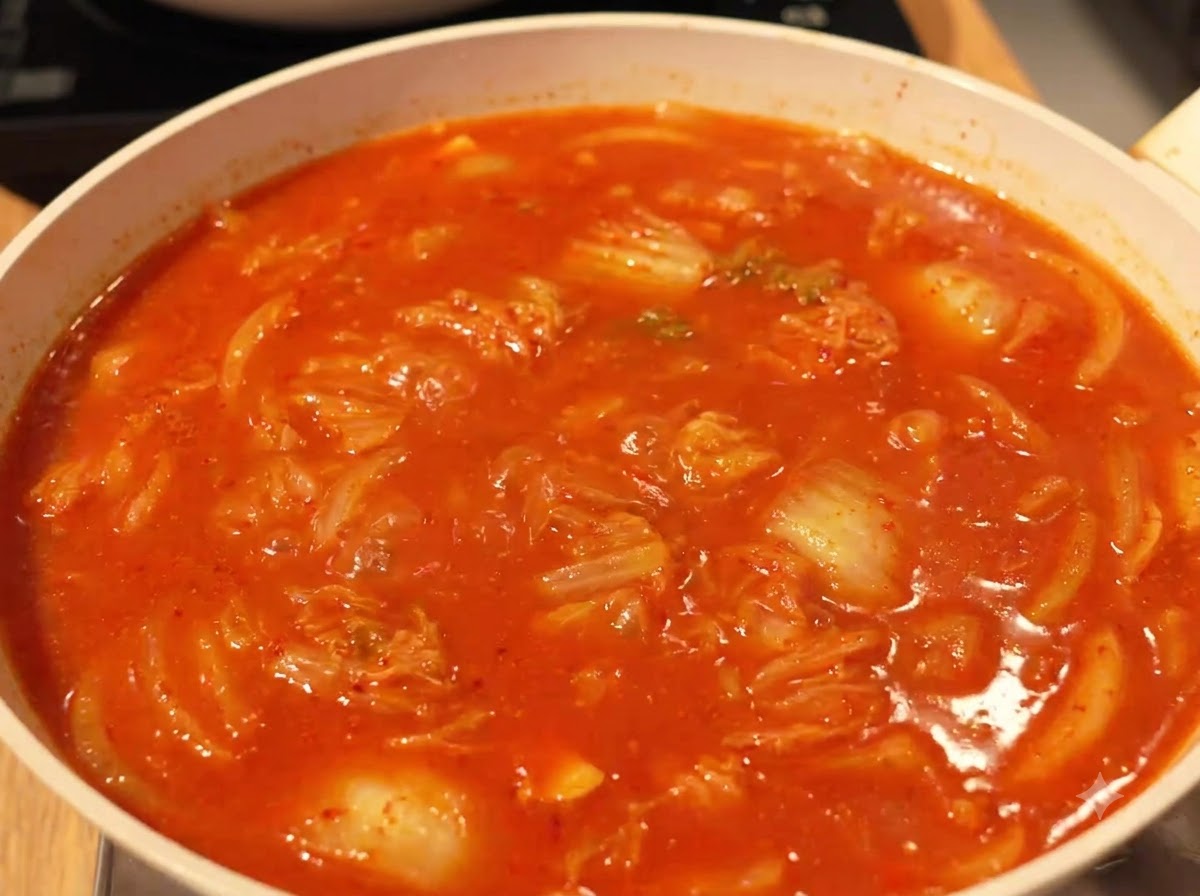 Un brodo ricco, rosso e ribollente che sobbolle con kimchi e ingredienti in una padella sul fuoco.