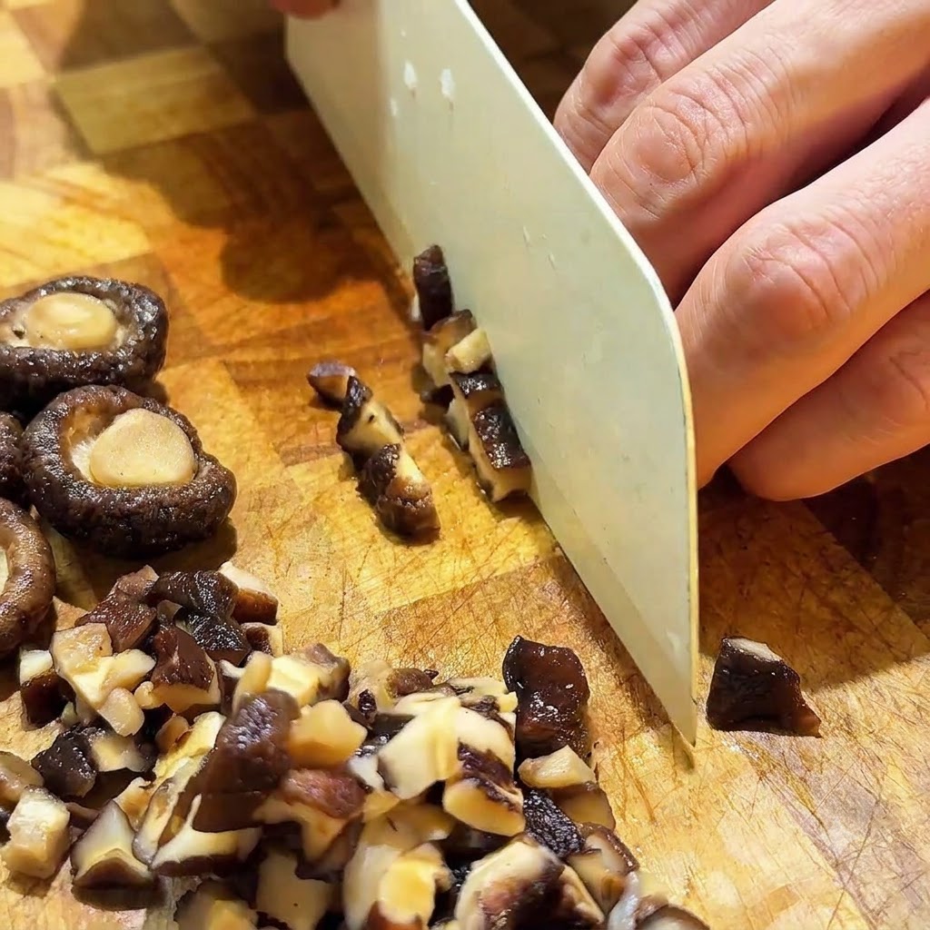 Funghi shiitake reidratati tagliati a piccoli cubetti con un coltello bianco su un tagliere di legno.