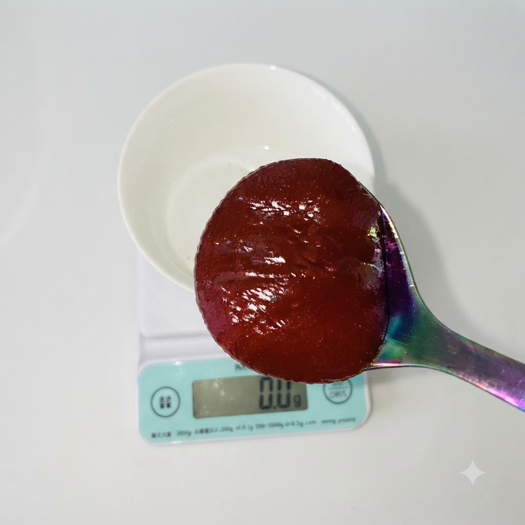 Un cucchiaio di pasta di peperoncino rossa coreana tenuto sopra una ciotola bianca poggiata su una bilancia da cucina digitale.
