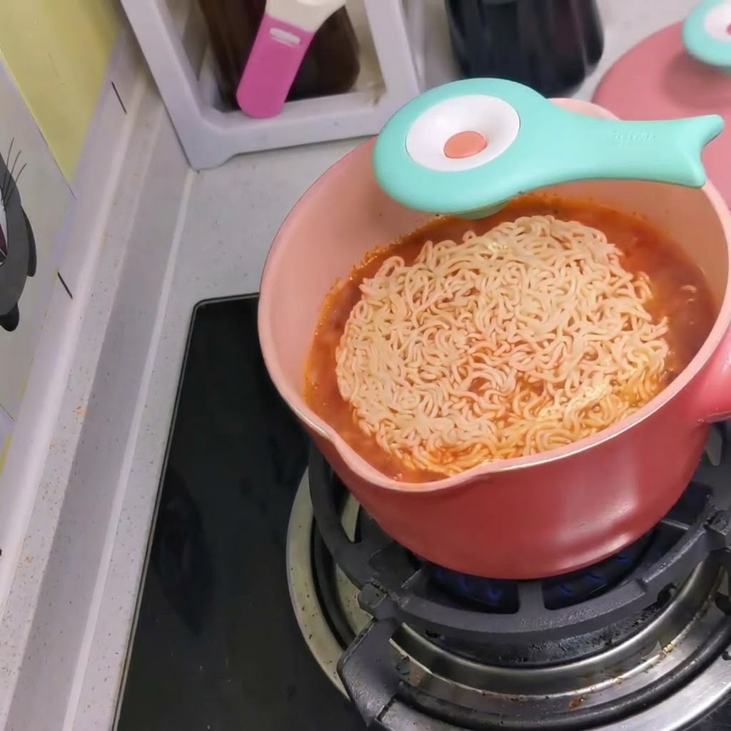 Una confezione di noodles istantanei gettata in una pentola rosa di brodo rosso piccante bollente su un fornello.