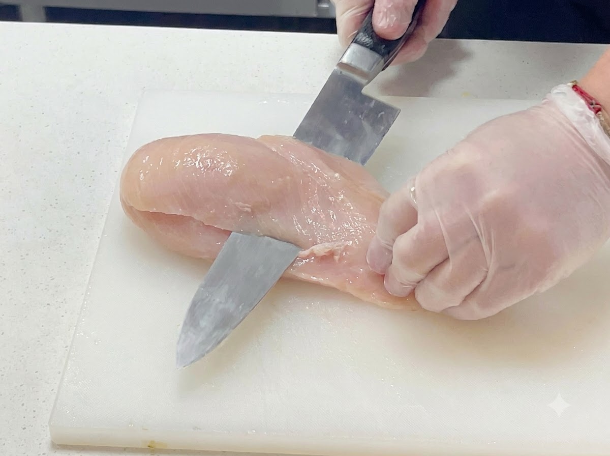Una vista ravvicinata di una mano guantata che taglia con cura un petto di pollo crudo orizzontalmente con un coltello da cucina su un tagliere bianco.