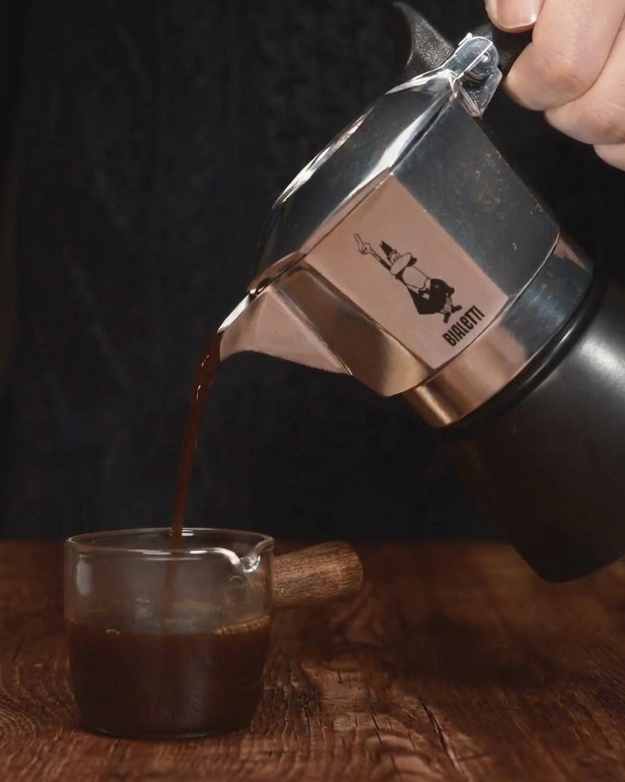 Espresso scuro versato da una moka dargento in un piccolo contenitore di vetro.