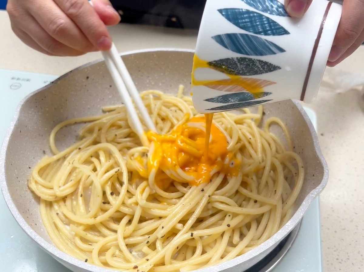 Denso composto di tuorlo duovo e formaggio versato da una ciotola sopra gli spaghetti cotti in padella mentre vengono mescolati rapidamente.