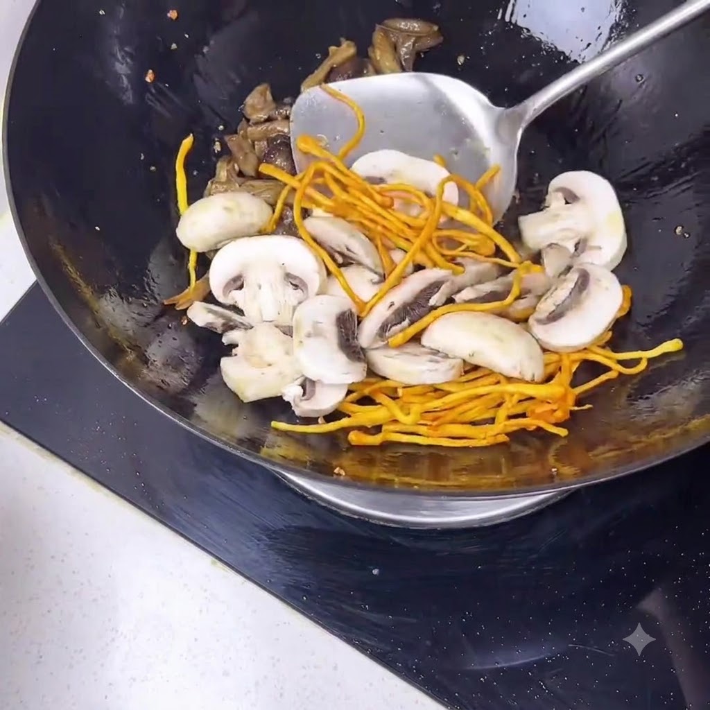 Una spatola di metallo che mescola funghi champignon affettati e fiori di cordyceps gialli allinterno di un wok nero.