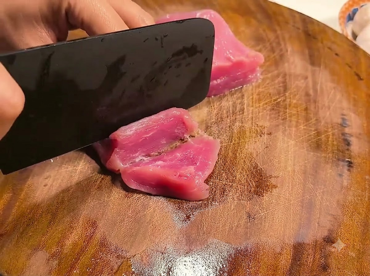 Mani che usano una mannaia per affettare il filetto di maiale rosa crudo su un tagliere.
